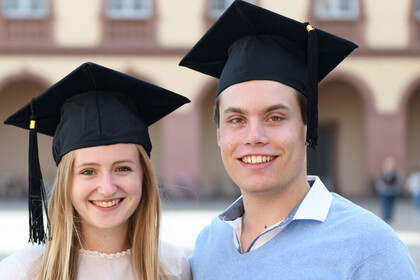Eine Bachelorabsolventin und ein Bachelorabsolvent stehen vor dem Mannheimer Schloss.