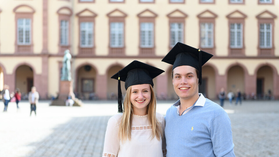 Eine Bachelorabsolventin und ein Bachelorabsolvent stehen vor dem Mannheimer Schloss.