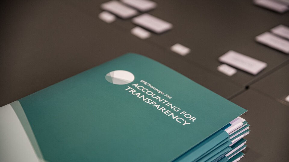 Türkisfarbene Mappen liegen auf einem schwarzen Tisch. Die Mappe haben die weiße Aufschrift "SFB/Transregio 266, Accounting for Transparency".