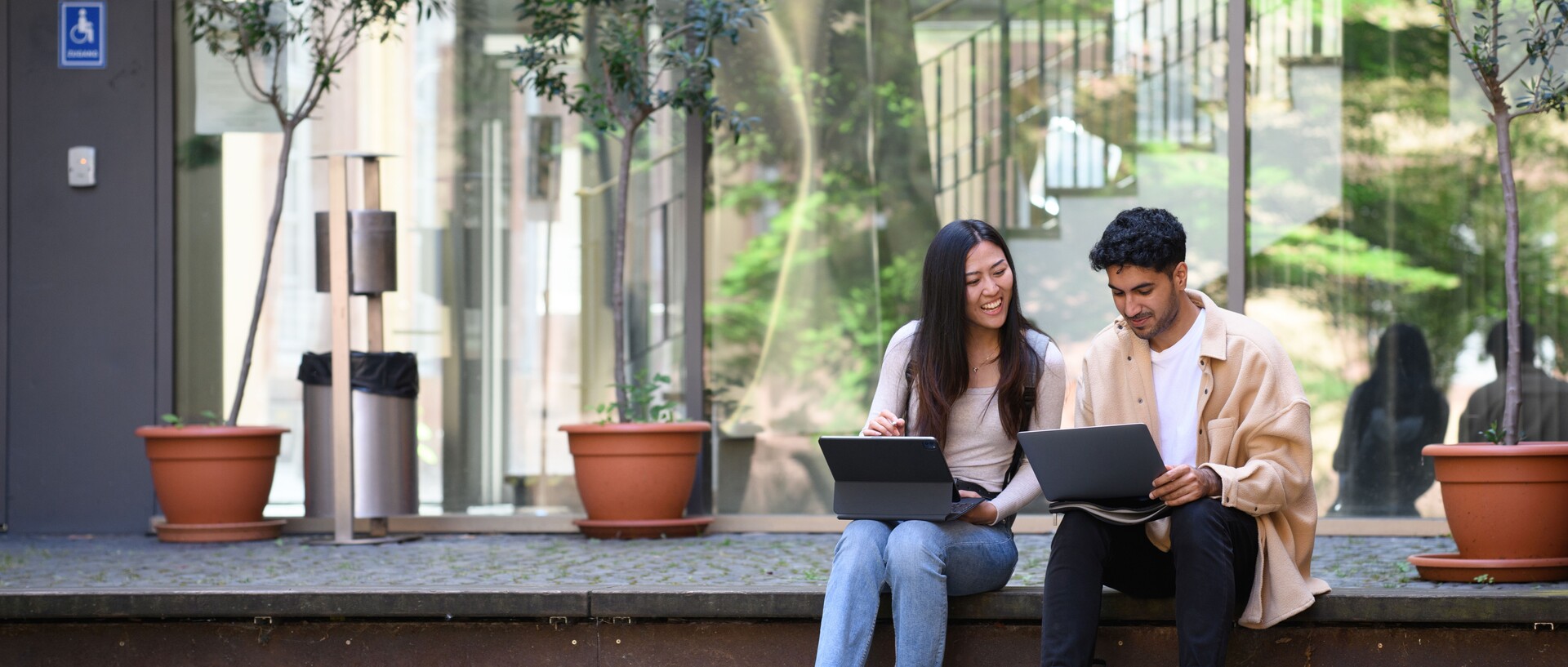 Eine Studentin und ein Student sitzen lachend auf einer Treppe und arbeiten an ihren Laptops.