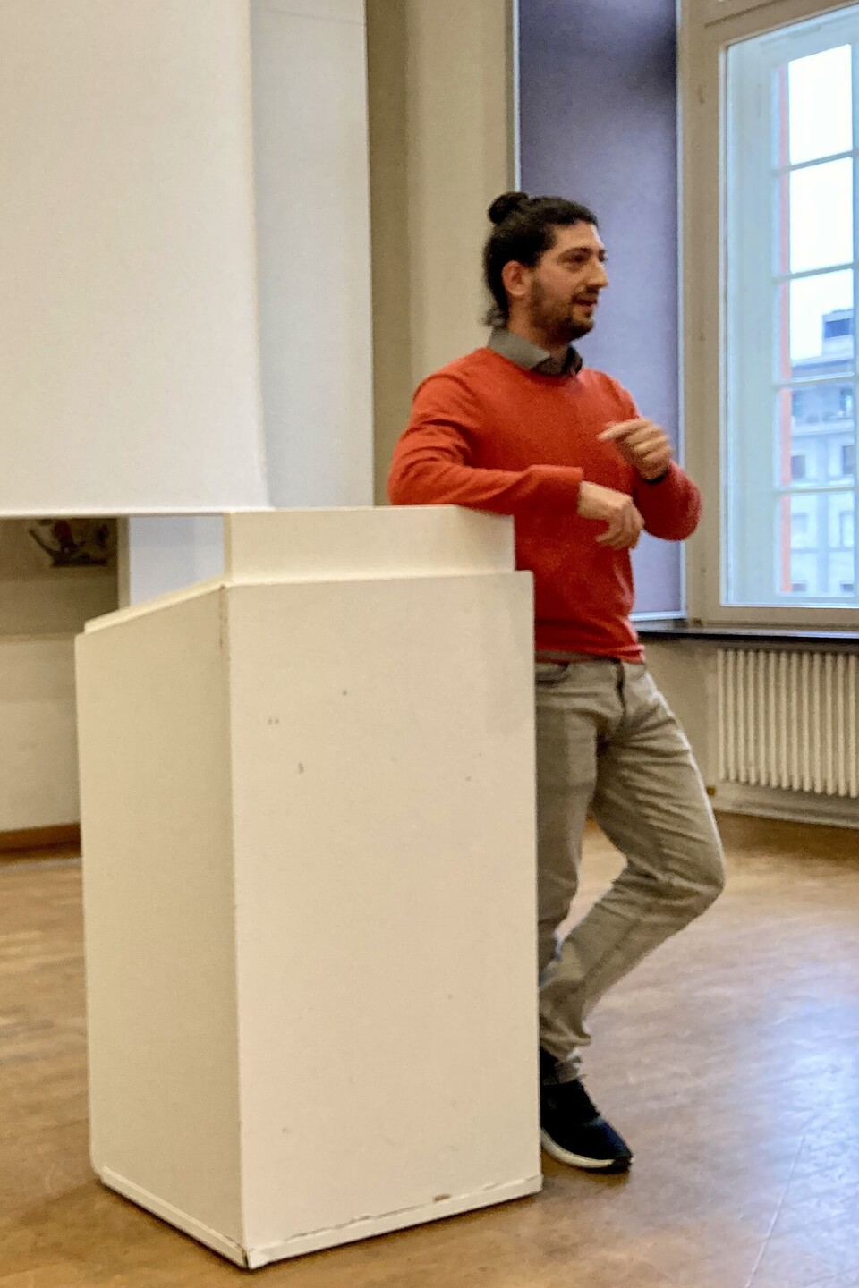 Prof. Mill lehnt am REdnerpult und spricht zum Publikum. Er trägt einen orangenen Pullover und hat lange Haare und einen Bart.