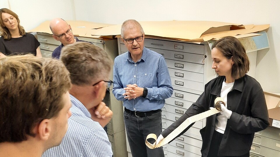 Die Teilnehmenden der Summer School stehen in einem Depotraum in einem Archiv. Eine Frau hält eine aufgerollte Pergamentrolle in der Hand, während andere ihr aufmerksam zuhören.