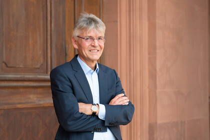 Professor Spengel trägt einen Anzug und hat die Arme ineinander verschränkt. Er steht vor einer Holztür.