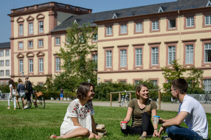 Zwei Studentinnen und ein Student sitzen auf einer grünen Wiese. Sie tragen frühlingshafte Kleidung und halten Kaffee-To-Go-Becher in ihren Händen.