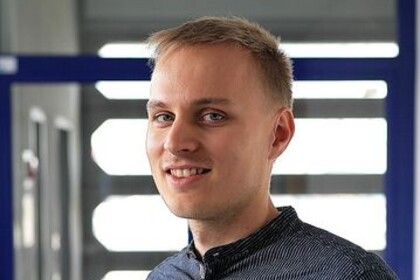 Portrait von Christoph Steinert. Er trägt ein dunkelgraues Hemd und steht lächelnd vor einer Fensterfront.