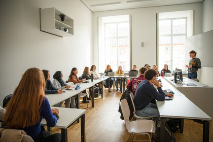 Studierende sitzen in einem Seminarraum an Tischen und schauen nach vorne zum Dozierenden.