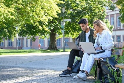 Zwei Studierende sitzen auf einer Bank auf dem Gelände der Universität Mannheim. Die Person links hat kurze schwarze Haare und trägt ein weißes T-Shirt mit grüner Jacke. Rechts daneben sitzt eine Person mit langen blonden Haaren. Sie trägt ein weißes Outfit. Beide haben Laptops auf ihren Schoß liegen.