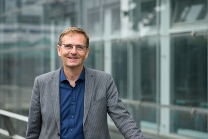 Portrait von Oliver Dickhäuser. Er trägt ein dunkelblaues Hemd sowie einen grauen Blazer und steht lächelnd vor einer Fensterfront.
