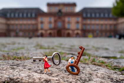 Zwei Miniaturfiguren, eine Frau im roten Kleid und ein Mann in blauer Latzhose, stehen auf Kopfsteinpflaster und tragen übergroße Schlüssel; im Hintergrund ist unscharf das Mannheimer Schloss zu erkennen.