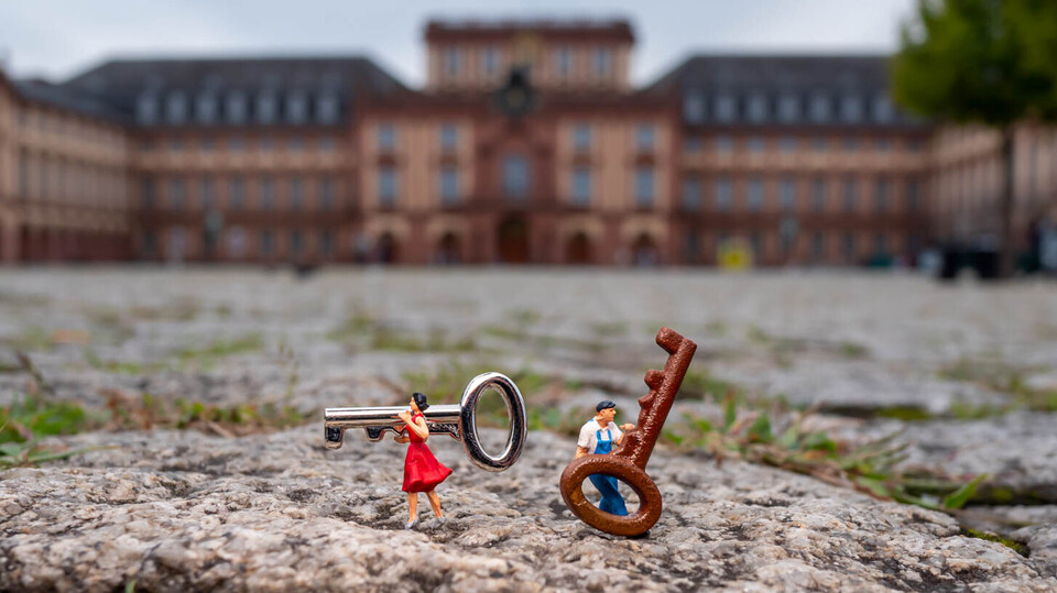 Zwei Miniaturfiguren, eine Frau im roten Kleid und ein Mann in blauer Latzhose, stehen auf Kopfsteinpflaster und tragen übergroße Schlüssel; im Hintergrund ist unscharf das Mannheimer Schloss zu erkennen.