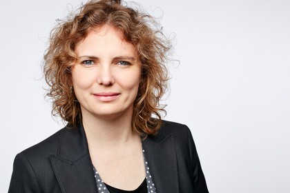 Portrait von Johanna Gereke. Sie trägt einen schwarzen Blazer und steht vor einer hellgrauen Wand.
