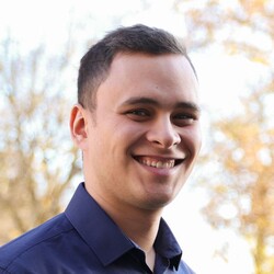 Dennis Uhrig hat dunkelbraune kurze Haare und braune Augen. Er trägt ein dunkelblaues Hemd und lächelt.