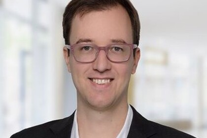 Portrait von Andreas Gulyas. Er trögt ein weißes Hemd sowie einen schwarzen Blazer und steht lächelnd in einem Gebäude.