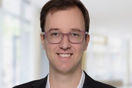 Portrait von Andreas Gulyas. Er trögt ein weißes Hemd sowie einen schwarzen Blazer und steht lächelnd in einem Gebäude.