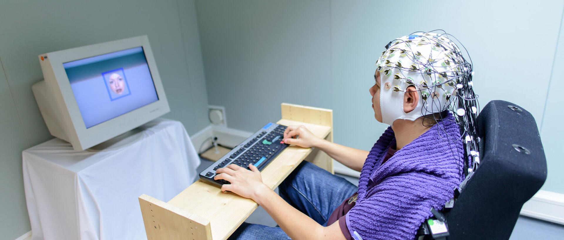 Eine Person trägt eine Haube mit Elektroden auf dem Kopf. Sie sitzt an einer Tastatur vor einem Monitor.