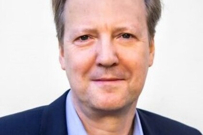 Portrait von Tom Krebs. Er trägt ein hellblaues Hemd sowie einen dunkelblauen Blazer.
