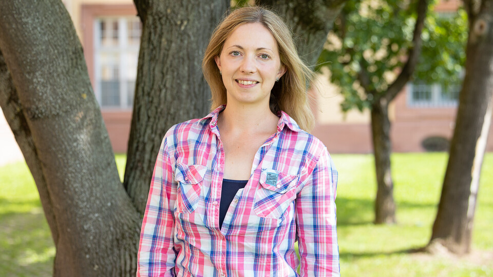 Britta Düvelmeyer trägt ein rosa-blau kariertes Hemd. Sie lächelt und steht vor einem Baum und einer grünen Wiese.