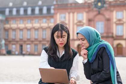 Zwei internationale Forscherinnen sitzen auf dem Ehrenhof vor dem Schloss und blicken auf einen Laptopbildschirm
