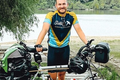Porträt von Joscha Friedrich in der Reihe Alumnus of the Month. Friedrich trägt Trikot und Radlerhose. Er steht an einem Flussufer und stützt sich auf sein Tourenrad. Damit ist er gerade auf dem Weg von Berlin nach Thailand. ️️Link: Instagram Alumnus of the Month⁠