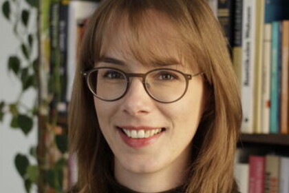 Portrait von Almuth Ebke. Sie trägt einen schwarzen Pullover und steht lächelnd vor einem Bücherregal.