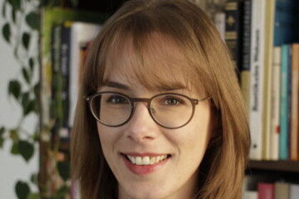 Portrait von Almuth Ebke. Sie trägt einen schwarzen Pullover und steht lächelnd vor einem Bücherregal.