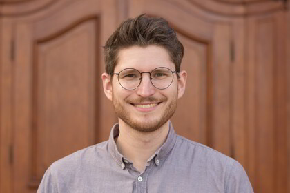 Portrait von David Izydorczyk. Er trägt ein graues Hemd und steht lächelnd vor einer braunen Holztüre.