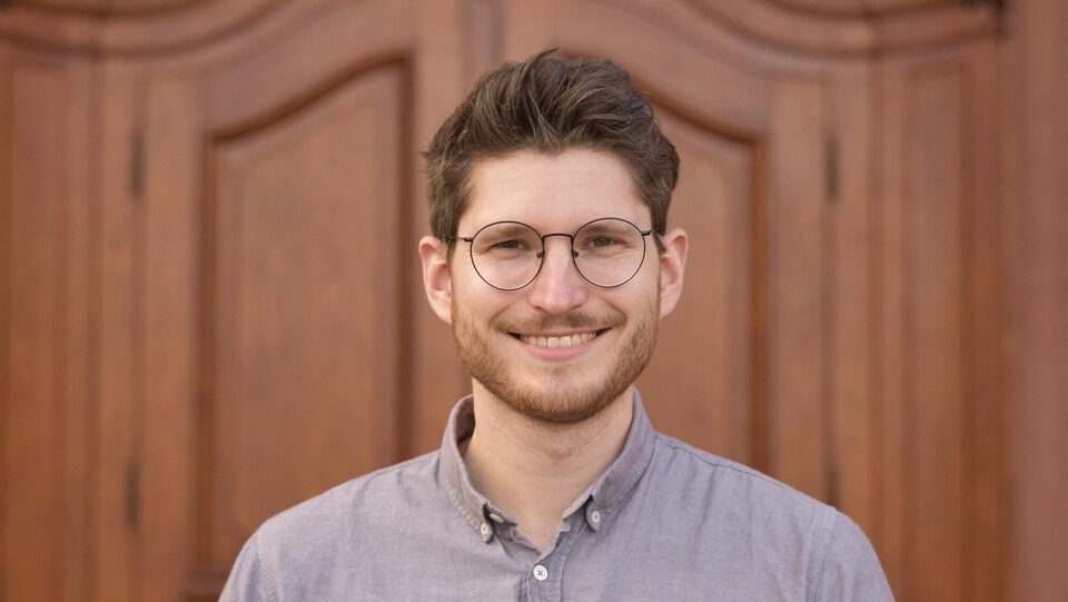 Portrait von David Izydorczyk. Er trägt ein graues Hemd und steht lächelnd vor einer braunen Holztüre.