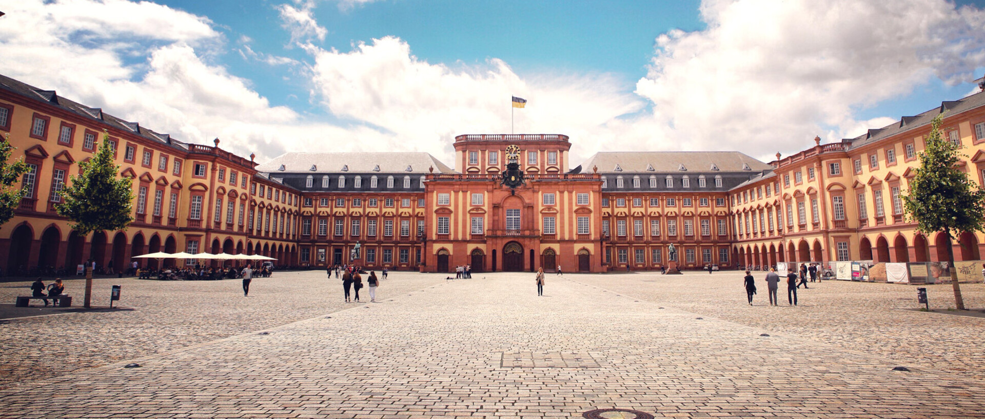 Das Barock-Schloss und der Ehrenhof der Universität Mannheim unter strahlend blauem Himmel. Das Schloss ist von unzähligen Fenstern, rotem Sandstein und einer gelben Fassade geprägt.