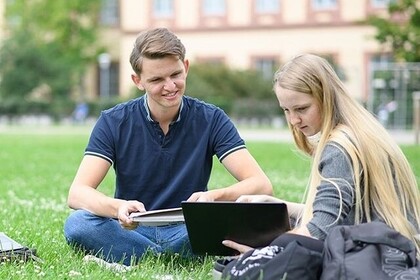 Zwei Studierende sitzen auf der Mensawiese der Universität Mannheim. Der Studierende links trägt ein blaues Oberteil und hat kurze braune Haare. Die Studentin rechts hat lange blonde Haare und trägt ein graues Oberteil. Beide blicken auf einen Laptop.