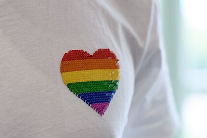 Ausschnitt vom Bild eines weißen T-Shirts. Über der linken Brust ist ein Paillettenherz in den Farben der Pride-Flagge aufgebügelt.