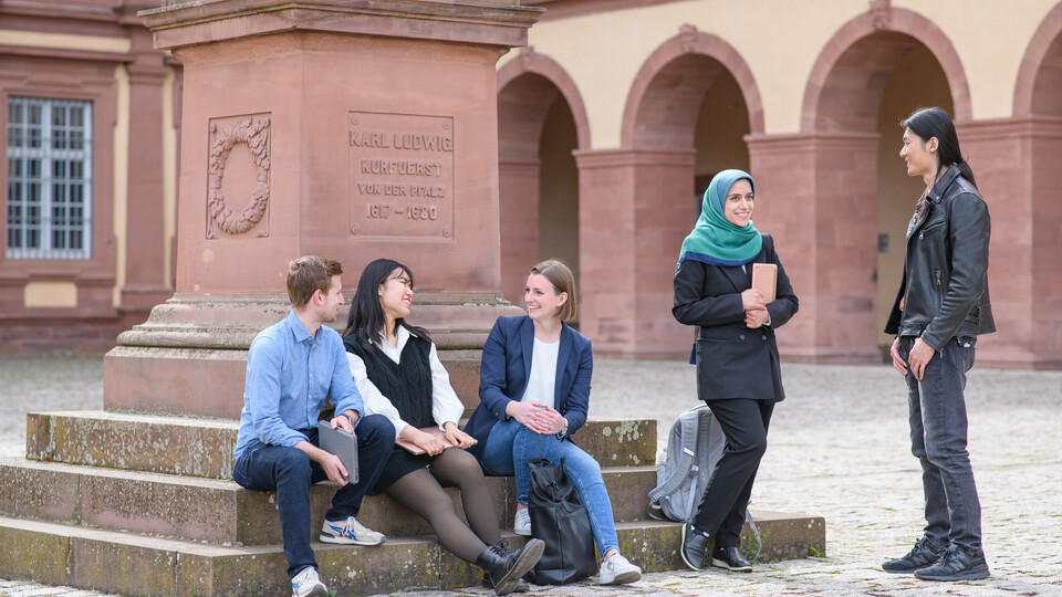 Drei Studierende sitzen auf der Karl-Ludwig-Statue an der Universität Mannheim, zwei stehen daneben im Gespräch.