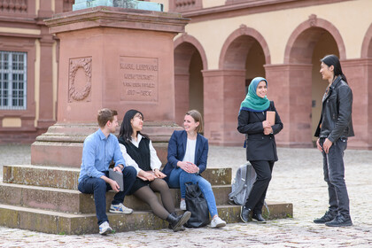Drei Studierende sitzen auf der Karl-Ludwig-Statue an der Universität Mannheim, zwei stehen daneben im Gespräch.