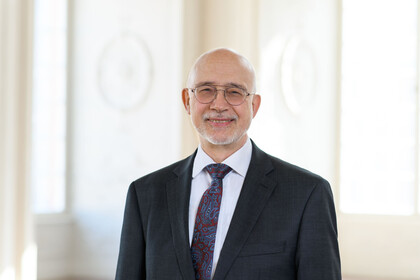 Portrait von Thomas Puhl. Er trägt ein weißes Hemd, eine rot-blaue Krawatte und einen schwarzen Blazer und steht lächelnd vor einer weißen Wand.