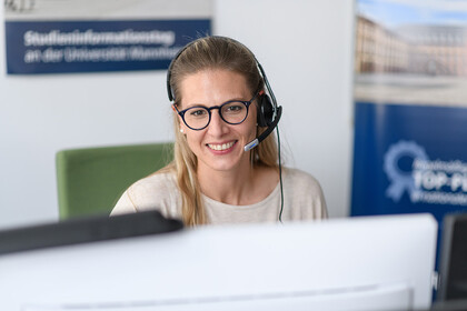 Eine Frau sitzt mit Kopfhörer in einem Büro, blickt in einen Bildschirm und lächelt.